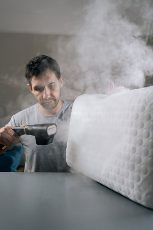 Serious man steam-cleaning white pillow using handheld steamer, removing allergens, dust mites and germs for deep-cleaned, fresh bedding and improved home hygiene.の写真素材