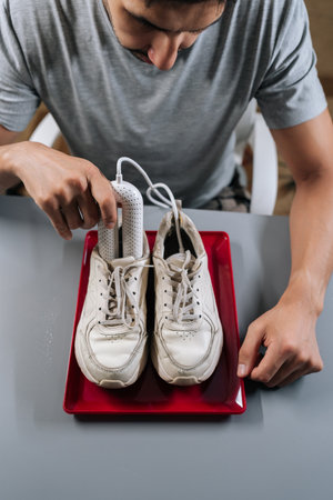 Top view of young adult man inserting portable electric dryer into white sneakers on red tray,...