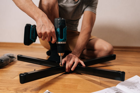 Man assembling new office chair base with electric screwdriver, constructing furniture at home on a wooden floor, following paper instructions for diy assembly project.の写真素材