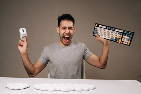 Excited young man shouts with open mouth and wide eyes, holding gaming keyboard in one hand and...