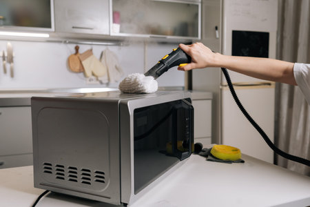 Closeup of cleaning service employee disinfecting microwave top with steam cleaner, removing grease and germs for spotless kitchen appliance. Concept of domestic hygiene and routine maintenance.の写真素材