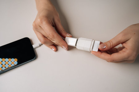 Close-up of female hands connecting mobile phone cable to white adapter while sitting at table,...