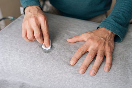 Person hands meticulously treating spill on gray fabric shirt using small white cloth soaked in...