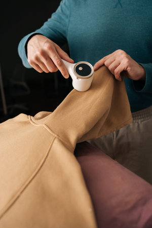 Vertical cropped shot of housekeeper using electric fabric shaver removing lint from beige sweatshirt sleeve for garment care and renewed clothing appearance, maintaining textile quality.の写真素材