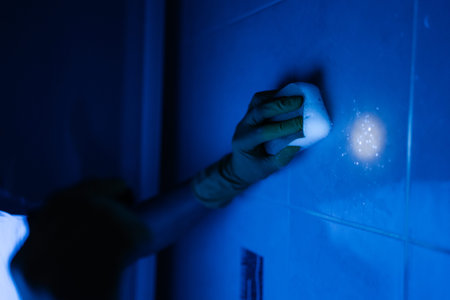 Hand in protective glove cleaning bathroom wall with sponge under ultraviolet light, visibly revealing glowing traces of hidden dirt and germs for thorough disinfection. Concept of household chores.の写真素材
