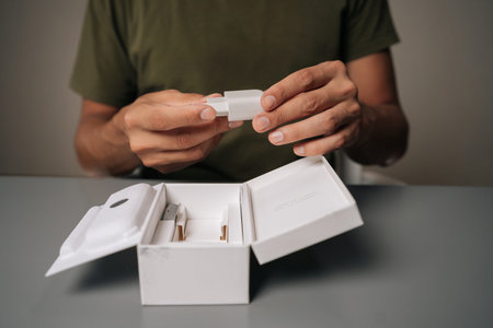 Close-up cropped shot of unrecognizable man unpacking box, holding white smartphone charger being unpacked from cardboard box, sitting on gray table. Concept of contemporary gadget unboxing.の写真素材