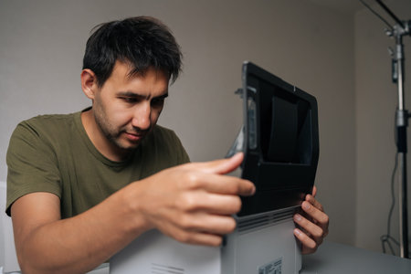 Pensive technician male examining components of disassembled laser printer, troubleshooting and performing maintenance in workshop environment, sitting at table on isolated background.の写真素材