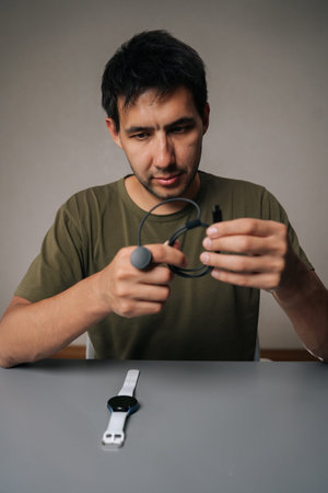 Vertical portrait of pensive young man untangling black charging cable for smart watch sitting at table with watch on isolated background, importance of keeping device powered up.の写真素材