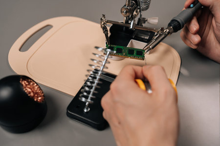 Close-up hand of technician soldering computer ram module using a helping hands tool and a soldering iron, repairing electronic components on a circuit board, working with microchips and technology.