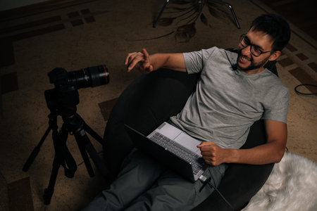 Happy young content creator in glasses smiling and pointing at camera while recording video blog or...