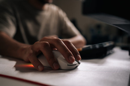Close-up of male hand using white rgb gaming mouse on white mousepad, with keyboard and monitor in...