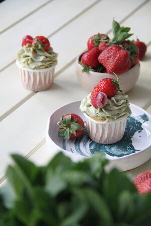Strawberry creamy cupcakes and green leaves, mint with flowers on white wooden kitchen background. Sweets for breakfest. Tasty food, beauty of delicious and art concept. Juicy colors, seasonal snack.の写真素材
