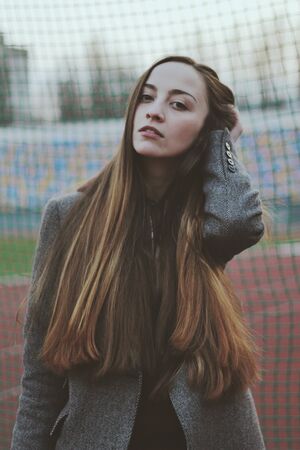 Sensual portrait of beautiful young girl walking in the old abandoned stadium on cold autumn day. Caucasian female model in front of grid. Human emotions, facial expression, fashion, beauty.の写真素材