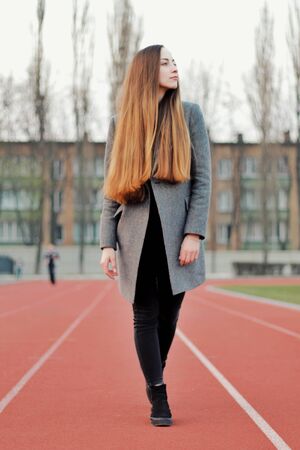 Sensual portrait of beautiful young girl walking in the old abandoned stadium on cold autumn day. Caucasian female model in front of city. Human emotions, facial expression, fashion, sport, lifestyle.の写真素材