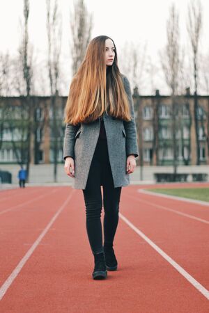 Sensual portrait of beautiful young girl walking in the old abandoned stadium on cold autumn day. Caucasian female model in front of city. Human emotions, facial expression, fashion, sport, lifestyle.の写真素材