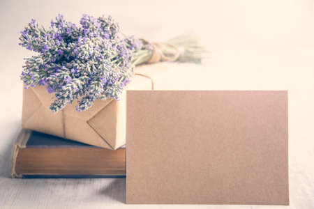 Empty greeting kraft card in front of a lavender bouquet, wrapped gift and old book over a white wood background. Vintage style.の写真素材