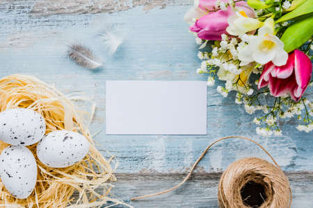 Top view of easter eggs in a nest. Spring flowers and feathers over blue rustic wood background. Empty card.の写真素材