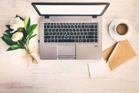 Top view of a desk with computer, coffee cup, kraft envelope, blank note and a bunch of peony flowers over white wood background.の写真素材