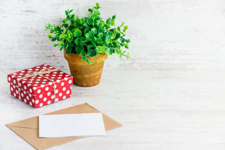 Red dotted gift box, empty card, kraft envelope and a green flower in a rustic ceramic pot. White wooden background, copy space.の写真素材