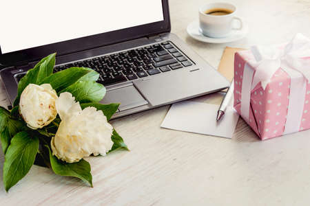 Side view of a deck with computer, bouquet of peonies flowers, cup of coffee, empty card and pink dotted gift box. White rustic wooden background. Romantic and feminine outlook.の写真素材