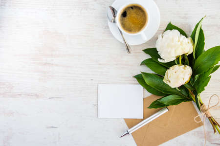 Top view of an empty greeting card, kraft envelope, peonies bouquet and a cup of coffee over white wooden rustic background.の写真素材