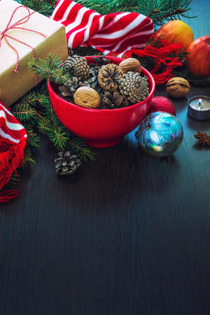 Christmas decoration - red bowl full of fir-cones, gift box wrapped in kraft paper, pine branches, candle, nuts, anise, apples, christmas toys and red and white striped winter scarf. Christmas theme. Dark wooden background.Copy spaceの写真素材