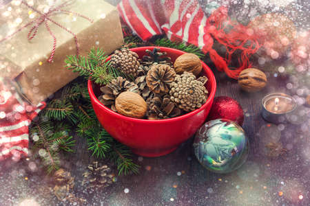 Christmas decoration - red bowl full of fir-cones, gift box wrapped in kraft paper, pine branches, candle, nuts, anise, apples, christmas toys and red and white striped winter scarf. Christmas theme. Dark wooden background.の写真素材