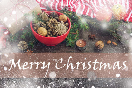 Christmas decoration - red bowl full of fir-cones, gift box wrapped in kraft paper, pine branches, candle, nuts, anise, apples and red and white striped winter scarf. Christmas theme. Merry Christmas text.の写真素材