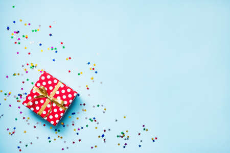 Top view of a red dotted gift box, scattered glittering star shaped confetti and colorful ribbons over blue background. Celebration concept. Copy space.の写真素材
