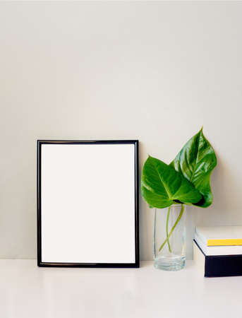 Black photo frame, green plant in a chrystal vase and a pile of books arranged against emty grey...