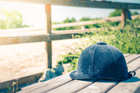 Close up of jockey's hat laid over wooden bench in front of horse stable fence.