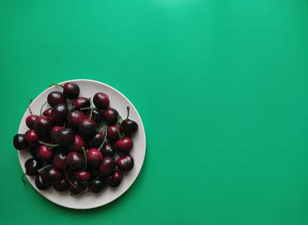 Overhead view of lots of dark red sweet cherries on white round plate on vibrant green backgroundの写真素材