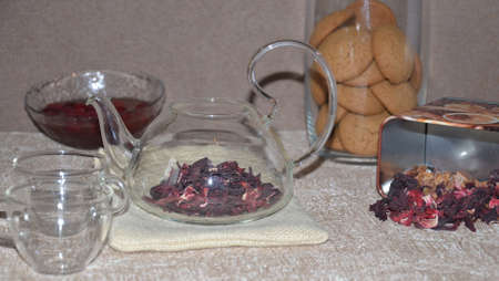 Serving table for breakfast, red tea in teapot. tea leaves, hibiscus and two glass cups for tea cookies on backgroundの写真素材