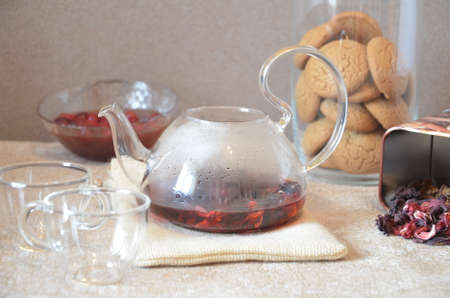 Serving table for breakfast, red tea in teapot. tea leaves, hibiscus and two glass cups for tea cookies on backgroundの写真素材