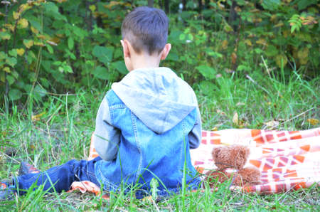 Back view of little boy sitting on the covering in autumn park, next to a toy bearの写真素材