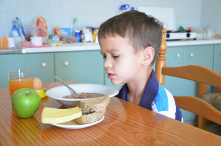 Cute boy eating a healthy delicious breakfast on the table in the kitchen. Diet. morning. Plate with porridge, egg on a stand, bread, cheese, green apple. Carrot peach orange tasty juice Useful and delicious foodの写真素材