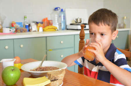 Cute boy drinking orange juice and eating muesli for breakfast. a healthy delicious breakfast on the table in the kitchen. Diet. morning. Plate with porridge, egg on a stand, bread, cheese, green apple. Carrot peach orange juice Useful and delicious foodの写真素材