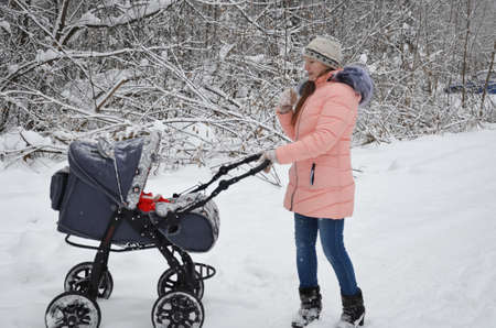 Young beautiful mother in a fur coat dancing while walking with a stroller in a city park at winter....