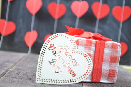 Gift or present box with red bow ribbon and a wooden heart with the inscription of a happy Valentine's day on wooden table for Valentines dayの写真素材
