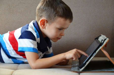 Little cute boy in a green T-shirt playing games on a tablet and watching cartoons. Toddler with tablet.の写真素材