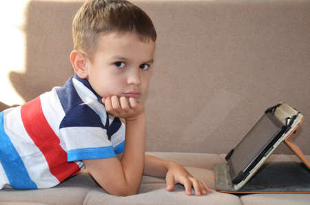 Tired young boy with glasses sitting and holding his head on the tablet in front of the computer, tablet and laptopの写真素材