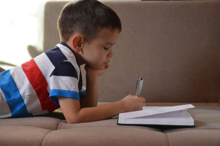 Close up little boy hand writing on paper, Kid writing on sofa in living room, student child boy holding colour pencil learning how to write homework at home, education conceptの写真素材