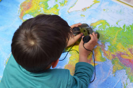 Kid is looking through binoculars around. Adventure and travel concept. Joyful background. Child is playing in his room in front of a map of the world.の写真素材