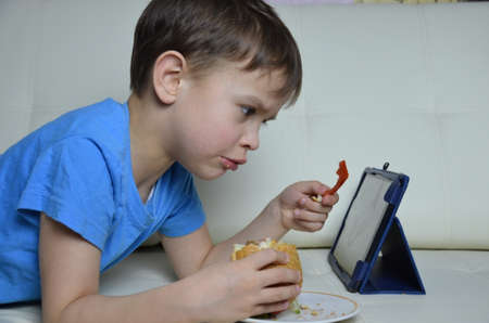 cute boy at home on the couch, eating a hamburger and looking at the tablet, watching cartoons or talking with friends he eat burgers and look at something on the tablet. he focused on something that is displayed on the screenの写真素材