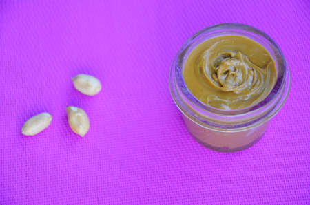 Jar and spoon of peanut butter and peanuts on purple background from top viewの写真素材