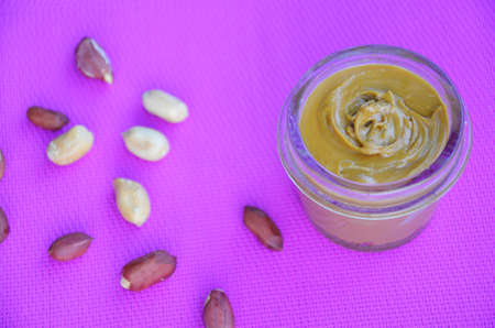 Jar and spoon of peanut butter and peanuts on purple background from top viewの写真素材