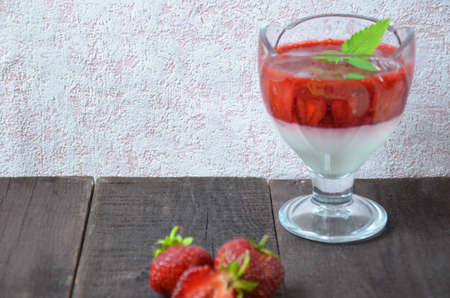 Summer dessert, panna cotta or yogurt or pudding or jam with strawberrieswith strawberries decorated with mint leaves. On a brown wooden table, bright sunlight. Copy space. close upの写真素材