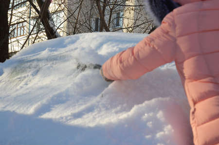 Winter portrait of a young woman cleaning snow from a car. Beauty blonde Model Girl laughs and cheerfully cleans the snow. Beautiful young woman outdoors.の写真素材