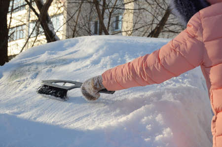 Winter portrait of a young woman cleaning snow from a car. Beauty blonde Model Girl laughs and...