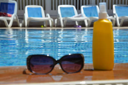 sunglasses and yellow sunscreen near a blue pool outside. concept of vacation, vacation, summer at...